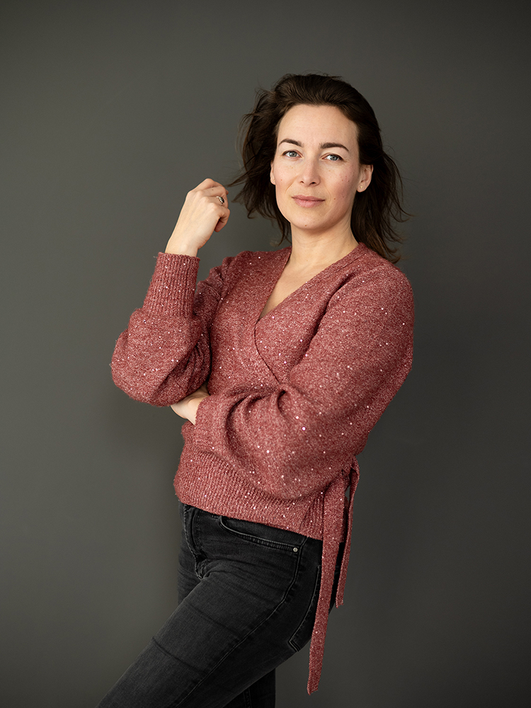 portretfoto van een vrouw met roze vest aan en donker haar die staat voor een grijze achtergrond